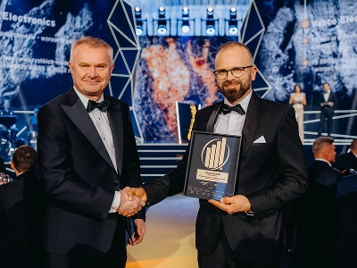 Et billede af to mænd i formel gallapåklædning, der giver hinanden hånden. Manden til højre, Maciej Góralski, holder en indrammet pris med et gyldent emblem, som viser, at det er prisen “Entrepreneur of the Year 2022”. I den slørede baggrund ses en scene med abstrakte geometriske strukturer og dynamiske blå og rød-orange grafiske mønstre. Flere gæster sidder ved borde i  eventhallen, der har dæmpet belysning.