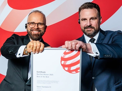 To mænd i formelt forretningstøj - den ene i smoking med briller, den anden i mørkeblåt jakkesæt - holder sammen et indrammet certifikat. Certifikatet viser tydeligt “Red Dot Winner 2025 Best of the Best” tildelt “Vasco Translator E1”. I baggrunden ses en stor grafisk installation med abstrakte røde og hvide bølgemønstre.