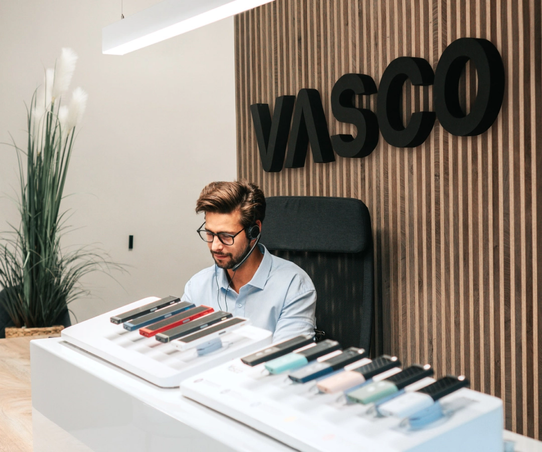 En mand fra kundeservice sidder ved et skrivebord og viser flere Vasco Translator-enheder i forskellige farver med et stort 'VASCO'-skilt på trævæggen bag sig.