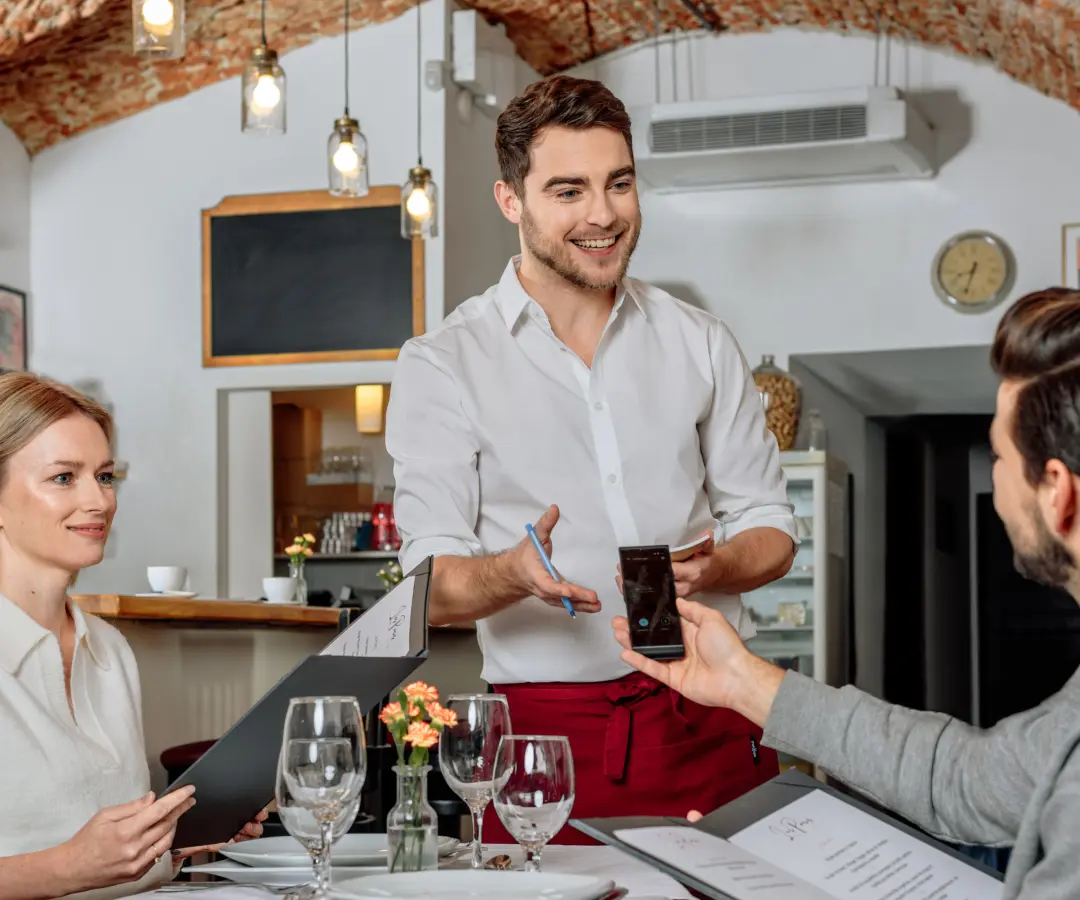 En smilende tjener står ved et restaurantbord, mens en siddende mandlig kunde holder en oversætter i hånden. En kvindelig kunde sidder i nærheden med et menukort i hånden. Bordet er dækket med glas, tallerkener og en lille blomstervase.