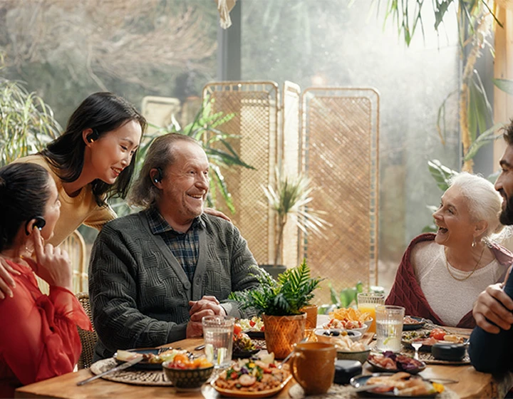 En familie, der sidder rundt om et bord med Vasco Translator E1-øretelefoner på, og som griner og snakker med hinanden.