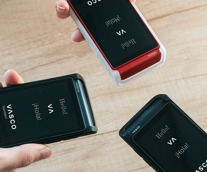 Tre personer holder Vasco Translator Q1-enheder og bringer dem sammen; skærmene viser startskærmen med Vasco-logoet og ordene “Hello!” og “¡Hola!”. Enhederne er vist i silikoneetuier i tre farver: Solid Black, Dark Caramel og Milky White.