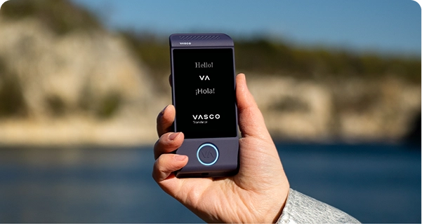 Et nærbillede af en hånd, der holder en lilla elektronisk Vasco-oversætter mod en lys udendørsbaggrund. Skærmen viser hilsenerne »Hello!«, »¡Hola!« og logoet »VASCO Translator«. Baggrunden viser et billede i blød fokus af et solrigt kystlandskab med blåt vand og lyse klipper under en klar himmel. Personen, der holder enheden, har en lysegrå eller sølvfarvet jakkeærme med struktur. 