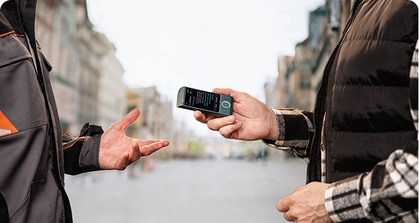 En person iført en sort, quiltet vest over en ternet skjorte holder en blågrøn Vasco-elektronisk oversætter vandret hen mod en anden person på en gade i byen. Den anden person, iført en grå og orange arbejdsjakke, rækker hånden frem, som om vedkommende er midt i en samtale. Enhedens skærm viser et oversættelsesinterface med tekst. I baggrunden ses et bredt, sløret billede af en gade i europæisk stil med høje bygninger under en lys, overskyet himmel. 