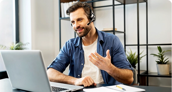 En venligt udseende mand med skæg og mørkt hår sidder ved et skrivebord iført et professionelt headset med mikrofon. Han smiler og gestikulerer med den ene hånd, mens han kigger på skærmen på sin bærbare computer; det ser ud til, at han er midt i et videomøde eller yder teknisk support. Han er klædt i en blå skjorte med knapper over en hvid T-shirt. På skrivebordet foran ham ligger en bærbar computer, en notesbog og en gul pen. Baggrunden er et lyst, moderne kontor med en minimalistisk sort metalhylde og grønne potteplanter. 